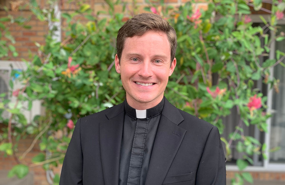 Smiling man in black clerical attire stands outdoors, vibrant green foliage with pink flowers in the background, conveying a cheerful mood.