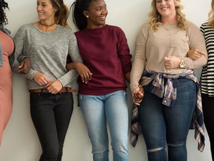 Five women stand arm-in-arm, smiling. They wear casual clothes in neutral tones. The background is plain, conveying a cheerful mood.
