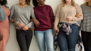 Five women stand arm-in-arm, smiling. They wear casual clothes in neutral tones. The background is plain, conveying a cheerful mood.