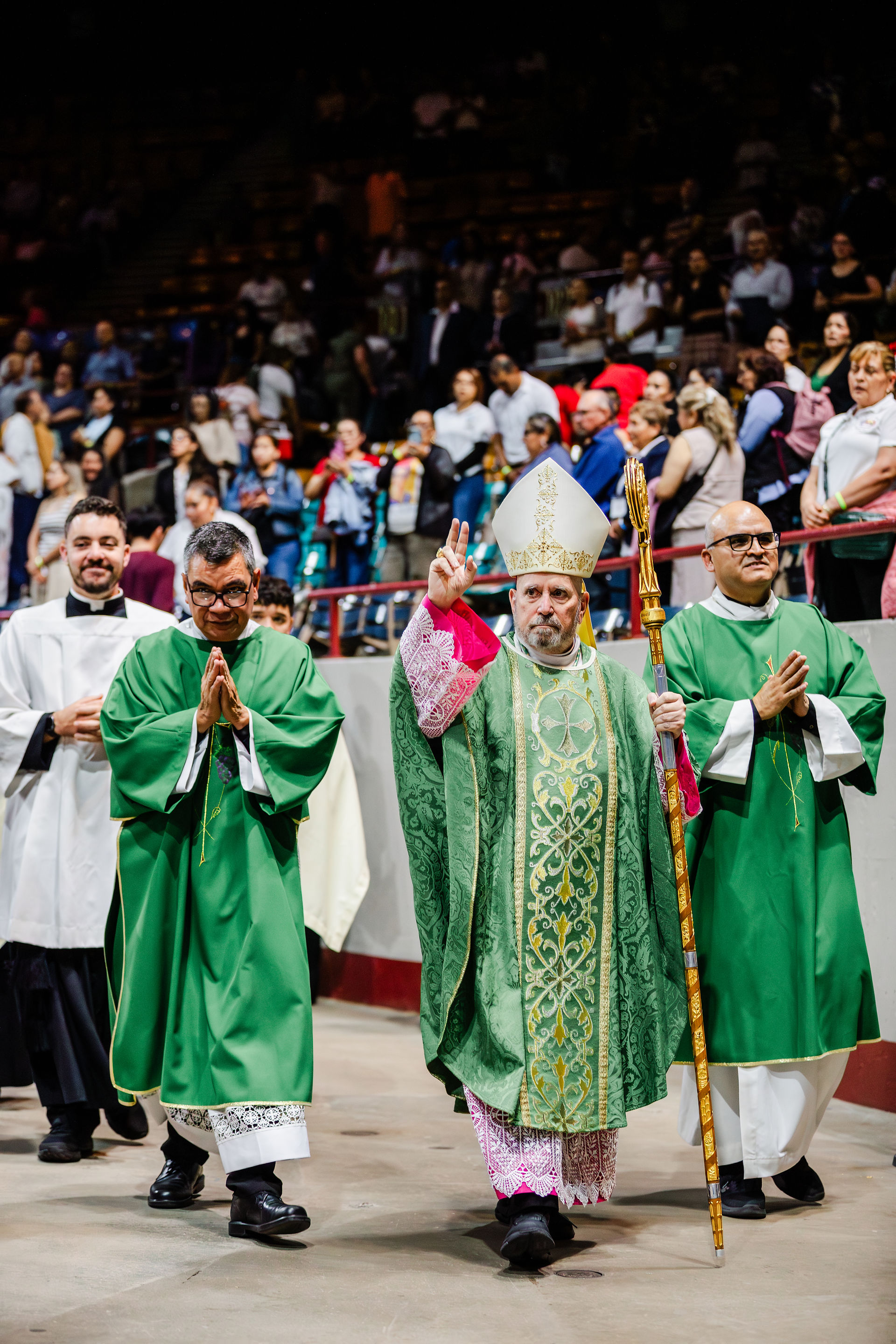 A religious procession led by a bishop wearing a mitre and holding a crosier, dressed in ornate green and gold vestments. He is flanked by clergy in green robes, with another figure in white robes behind. The background shows a crowd of attendees in an indoor venue, likely a church or hall, observing the ceremony. The image highlights traditional clerical attire and ritual.