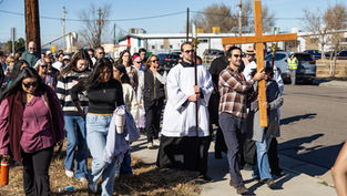 A group of people, including a person in a robe, walk down a street carrying a large wooden cross. The sky is clear and sunny.