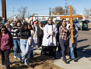 A group of people, including a person in a robe, walk down a street carrying a large wooden cross. The sky is clear and sunny.
