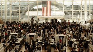 Busy airport security checkpoint with crowds navigating through scanners. Large American flag hangs in the background, creating a bustling scene.