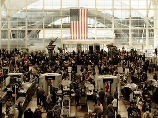 Busy airport security checkpoint with crowds navigating through scanners. Large American flag hangs in the background, creating a bustling scene.