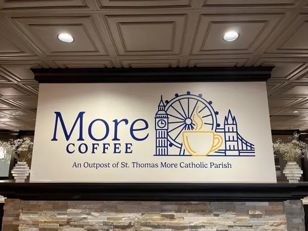 Wall sign in a coffee shop with a London skyline graphic and text: "More Coffee: An Outpost of St. Thomas More Catholic Parish."