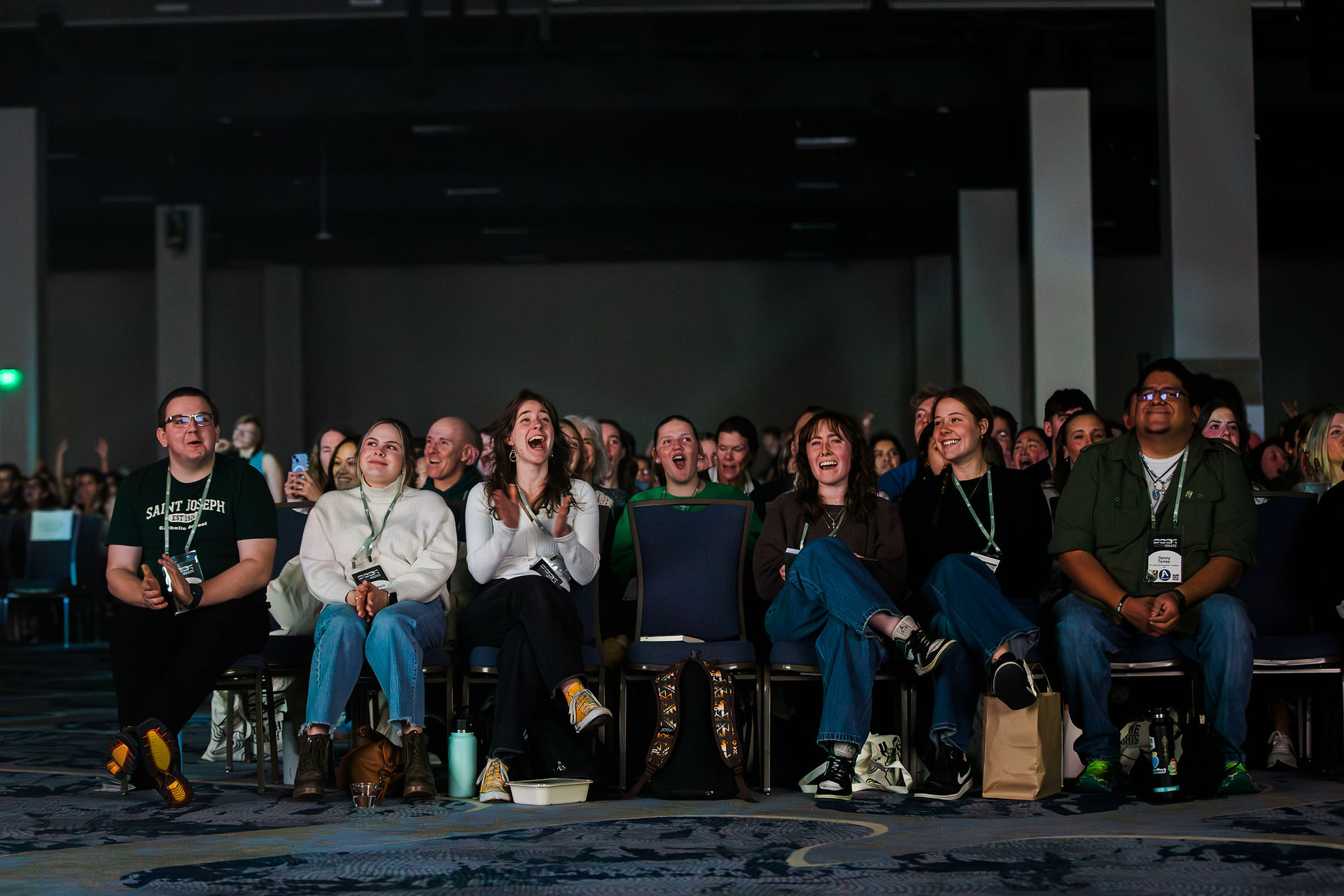 A group of people seated in a dimly lit room, smiling and clapping. They're watching something with excitement. Text on a shirt reads "SAINT JOSEPH."
