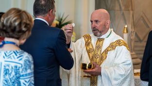 A bishop in white robes distributes communion during a religious service. Floral arrangements can be seen in the background. The mood is solemn, peaceful and prayerful.