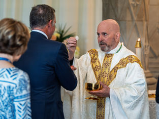 A bishop in white robes distributes communion during a religious service. Floral arrangements can be seen in the background. The mood is solemn, peaceful and prayerful.