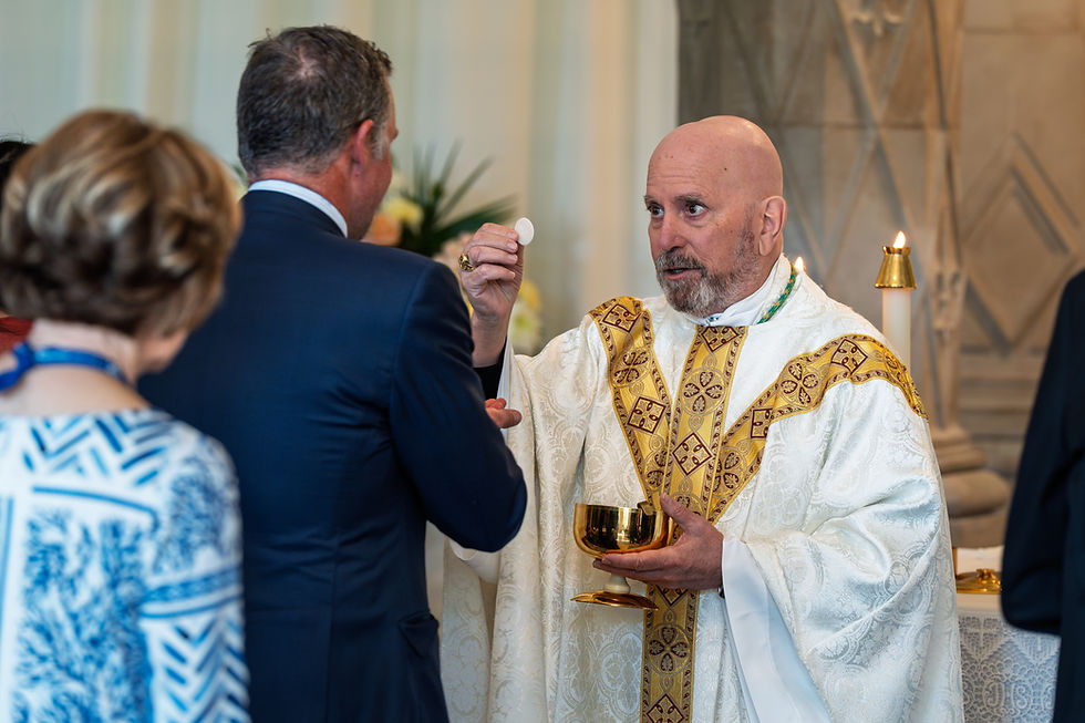 A bishop in white robes distributes communion during a religious service. Floral arrangements can be seen in the background. The mood is solemn, peaceful and prayerful.
