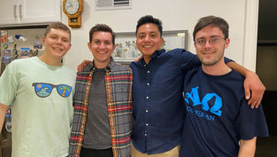 Four smiling friends pose indoors. One wears a light shirt with sunglasses print, another a plaid shirt, and two wear dark shirts with logos.