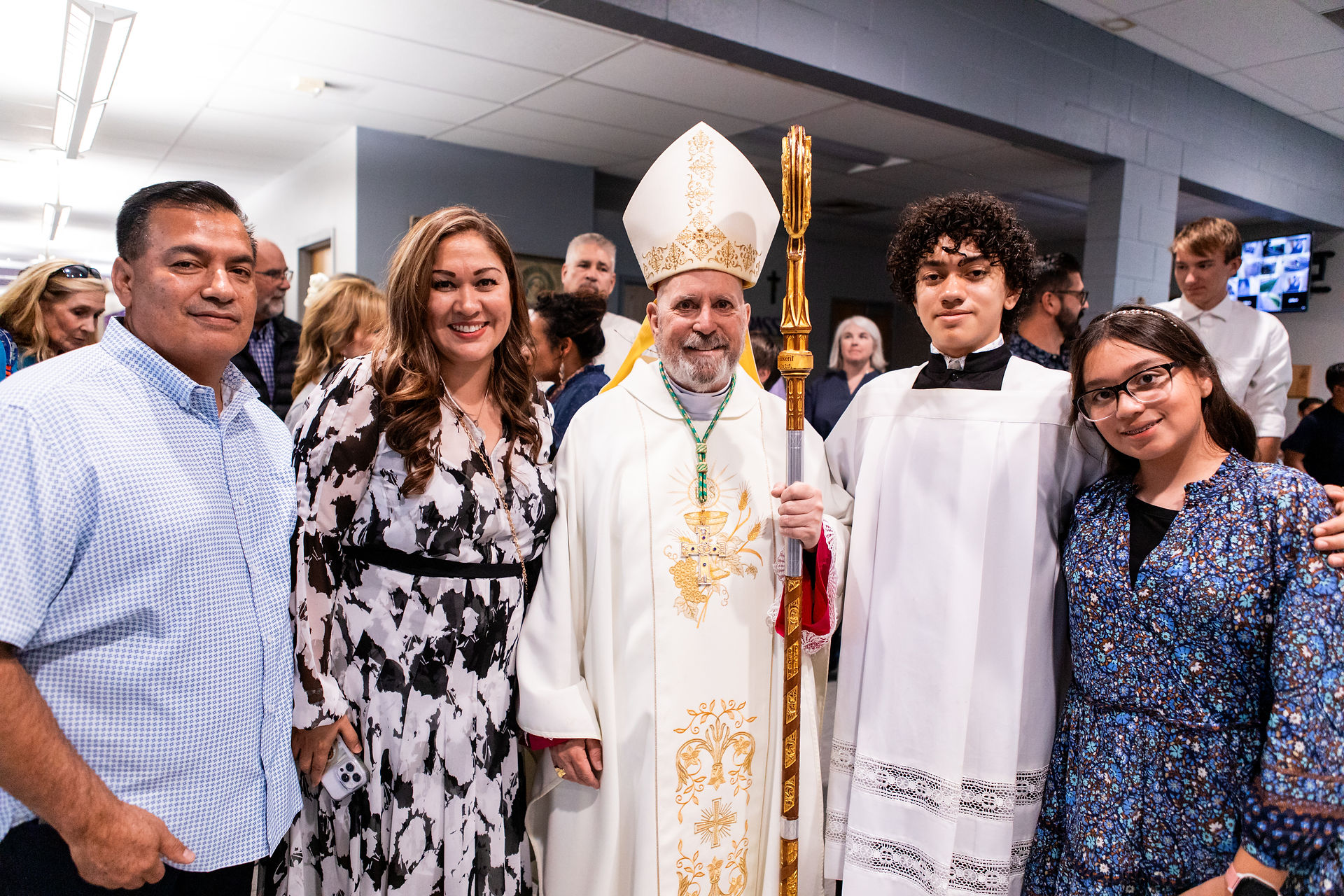 A group of five people, including a bishop in ornate robes, posing indoors. The atmosphere is joyful, with a crowd in the background.