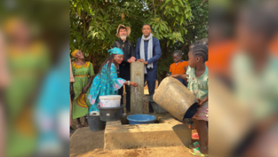 People and children gather at an outdoor hand pump, filling buckets and washing hands beneath leafy trees.