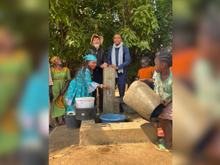 People and children gather at an outdoor hand pump, filling buckets and washing hands beneath leafy trees.