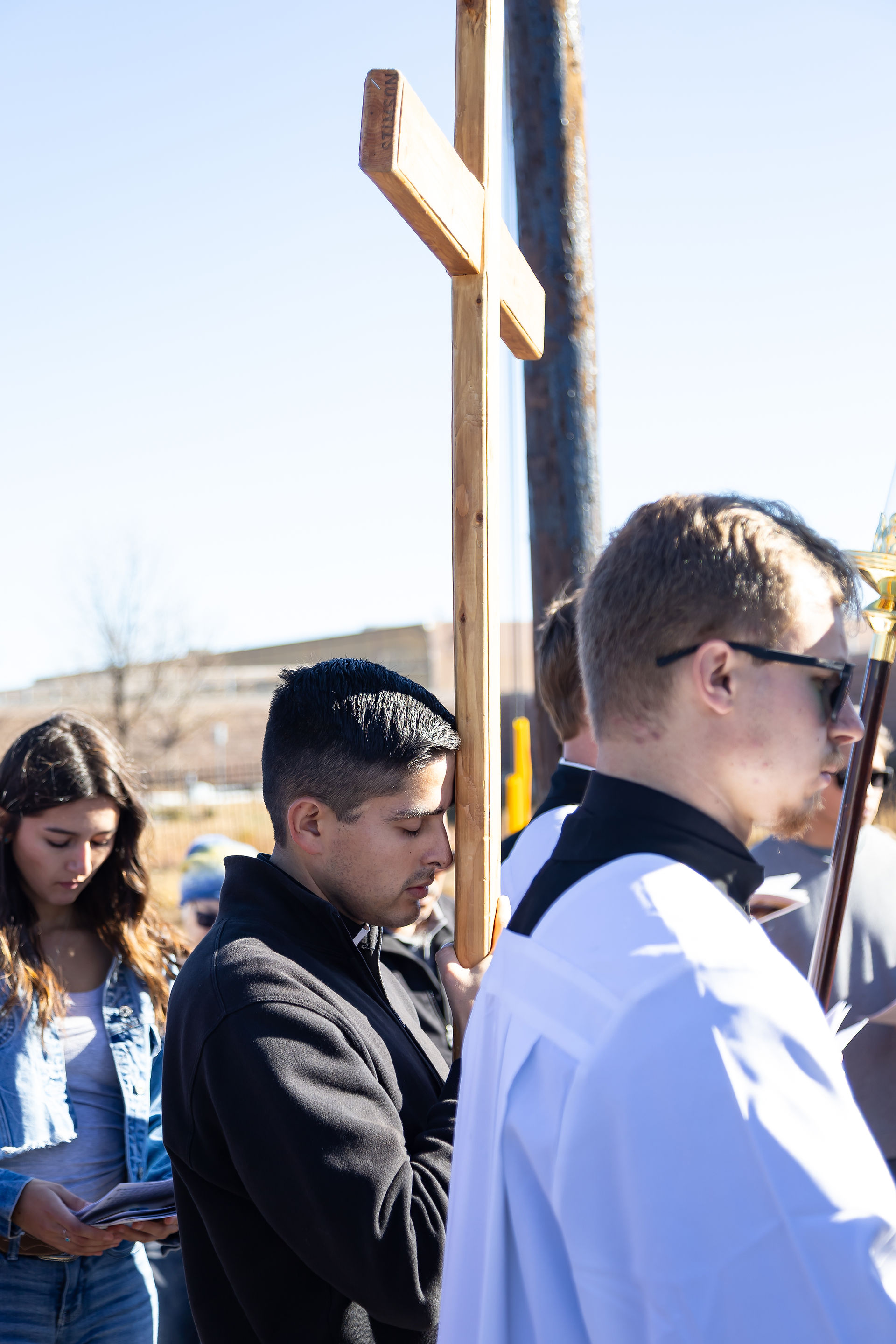 People participate in an outdoor religious procession, one man holding a wooden cross, others in prayer or reading. Sunny day, clear sky.