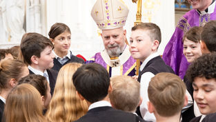 Bishop in purple vestments and mitre speaks with a group of schoolchildren gathered around him inside a church.