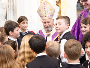 Bishop in purple vestments and mitre speaks with a group of schoolchildren gathered around him inside a church.