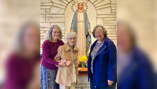 Three women smile in front of a statue of Mary in a stone alcove. The central woman holds hands with one on the left. Yellow flowers in front.