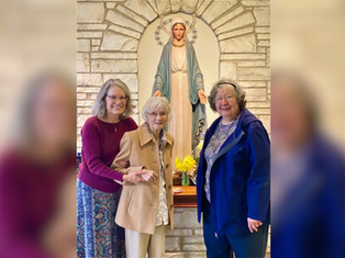 Three women smile in front of a statue of Mary in a stone alcove. The central woman holds hands with one on the left. Yellow flowers in front.