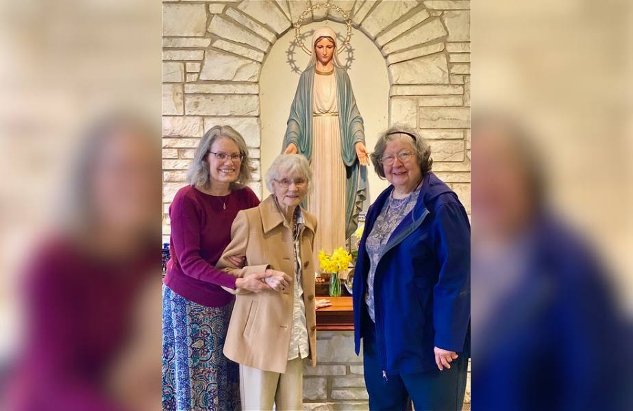Three women smile in front of a statue of Mary in a stone alcove. The central woman holds hands with one on the left. Yellow flowers in front.