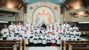 A large group of altar servers and priests, dressed in white and red robes, pose inside a church with a mural of Jesus on the wall behind.
