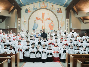 A large group of altar servers and priests, dressed in white and red robes, pose inside a church with a mural of Jesus on the wall behind.