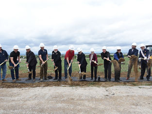 A group of people in hard hats ceremonially shovel dirt at a groundbreaking event on a grassy field, cloudy sky in the background.