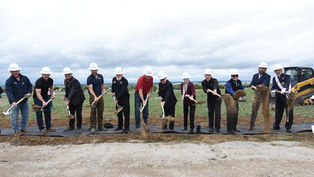 A group of people in hard hats ceremonially shovel dirt at a groundbreaking event on a grassy field, cloudy sky in the background.