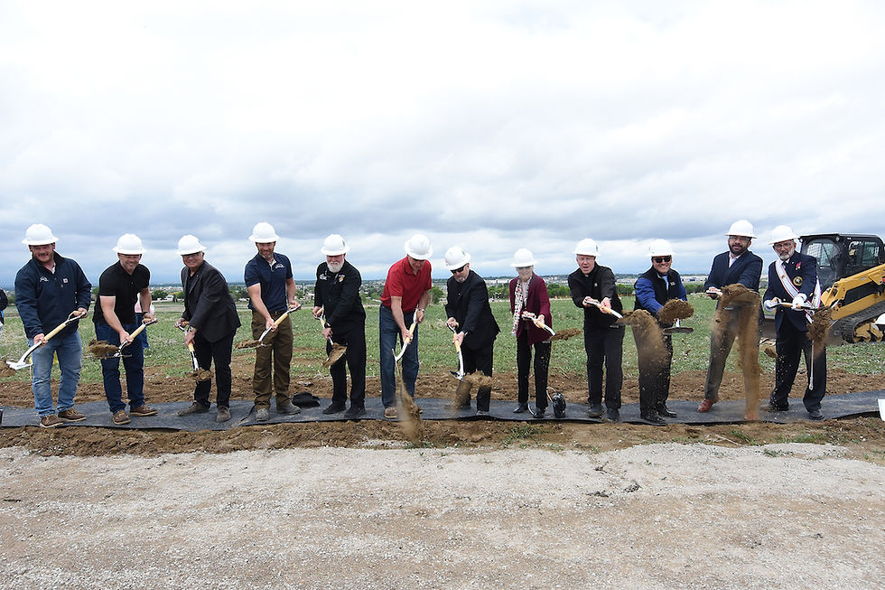 A group of people in hard hats ceremonially shovel dirt at a groundbreaking event on a grassy field, cloudy sky in the background.