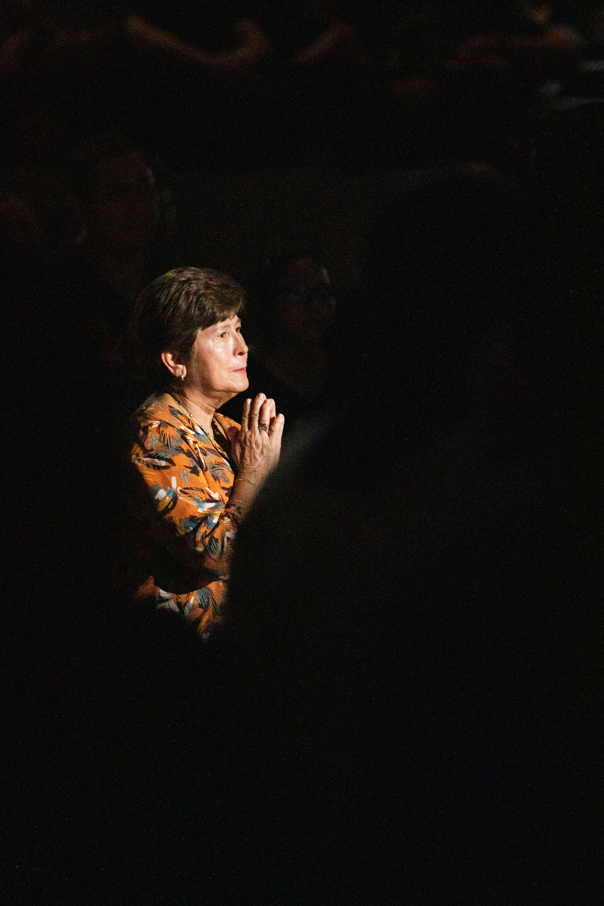 A woman in a floral shirt is clasping her hands in a dimly lit setting, focused intently. The background is dark, emphasizing her expression.