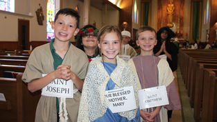 Children in costumes, labeled as saints, smile in a church with stained glass windows. Joyful atmosphere with vibrant colors and warm lighting.