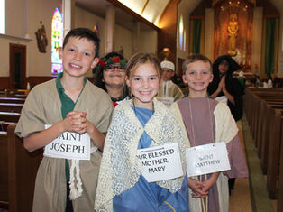 Children in costumes, labeled as saints, smile in a church with stained glass windows. Joyful atmosphere with vibrant colors and warm lighting.