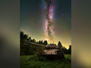 Rusty tank in a field under the Milky Way. Star-filled night sky creates a serene, contrasting backdrop. Trees silhouette the horizon.