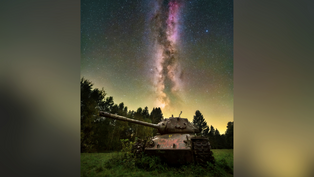 Rusty tank in a field under the Milky Way. Star-filled night sky creates a serene, contrasting backdrop. Trees silhouette the horizon.