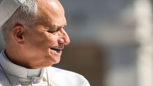 Smiling pope in white robes and skullcap, seen in profile against a blurred gray background, sunlit and peaceful