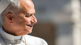 Smiling pope in white robes and skullcap, seen in profile against a blurred gray background, sunlit and peaceful