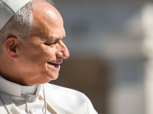 Smiling pope in white robes and skullcap, seen in profile against a blurred gray background, sunlit and peaceful