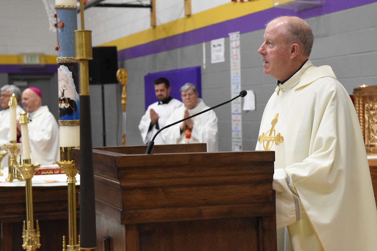 Priest in white robes speaking at a podium in a church. Clergy members in background. Candles on altar; purple and yellow walls.