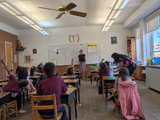Classroom scene with teacher and students. Children raise hands to answer. World map on whiteboard, natural light from windows, lively mood.