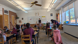 Classroom scene with teacher and students. Children raise hands to answer. World map on whiteboard, natural light from windows, lively mood.