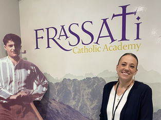 Woman smiling next to a cardboard cutout of a man. Background shows "FRASSATI Catholic Academy" text, with mountain imagery. Calm mood.