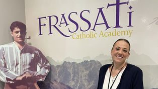 Woman smiling next to a cardboard cutout of a man. Background shows "FRASSATI Catholic Academy" text, with mountain imagery. Calm mood.