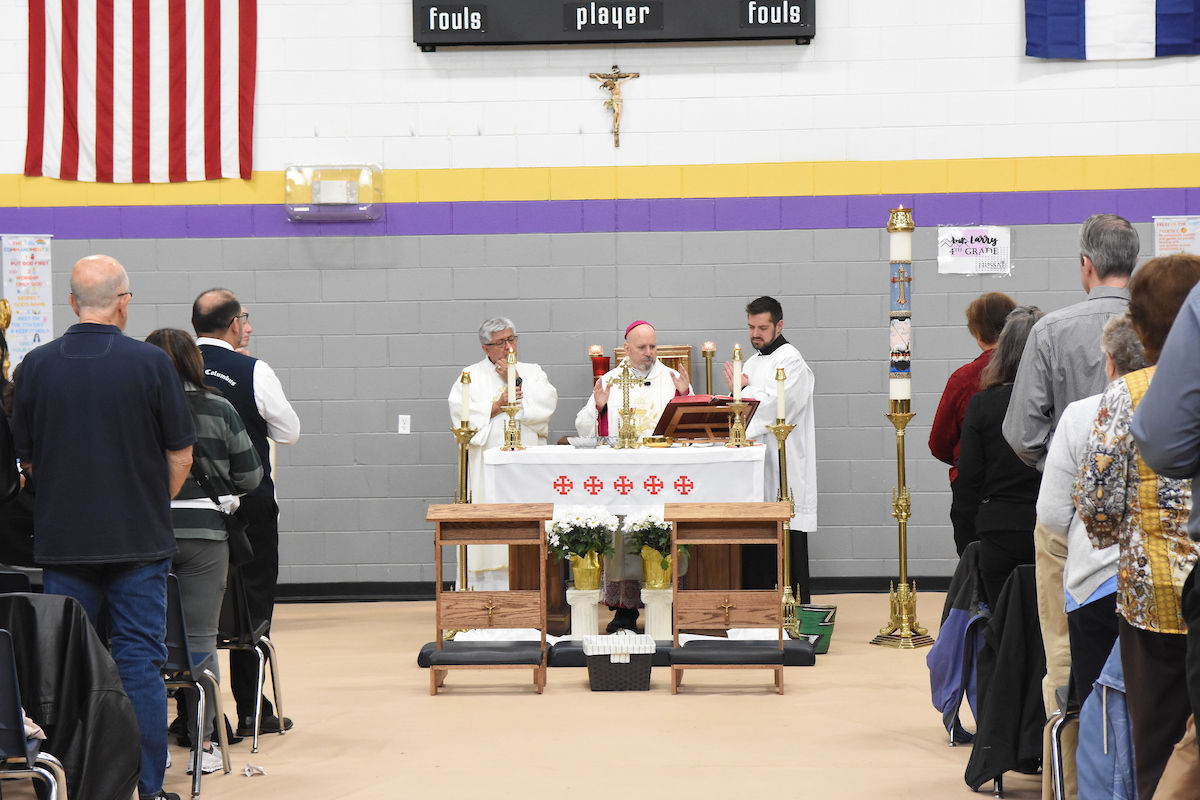 Priests conducting a mass in a gym with an altar and congregation. American flag on left, crucifix above, and gray walls in background.