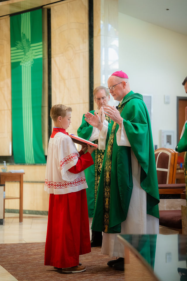 Bishop in green vestments blesses a boy in a red-and-white robe holding a book inside a church, solemn and ceremonial.