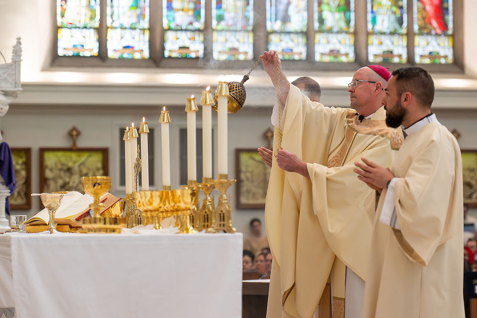 A bishop and clergy perform a ritual with incense in a church. Candles and gold chalices adorn the altar. Stained glass windows light the scene.