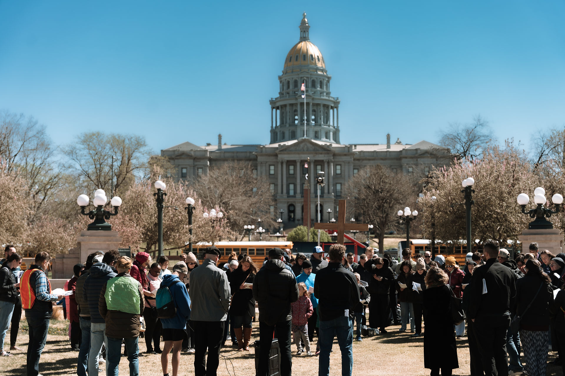 A group gathers in prayer on a lawn in front of the Colorado State Capitol, with a wooden cross at the center.