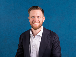 Smiling man in a suit and white shirt against a textured blue background. He has short hair and a beard. Mood is professional and friendly.