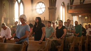 People standing and praying in a sunlit church with stained-glass windows. The mood is serene, with soft light highlighting wooden pews.