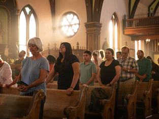 People standing and praying in a sunlit church with stained-glass windows. The mood is serene, with soft light highlighting wooden pews.