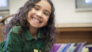 Smiling child with curly hair in a green shirt leans over a table with labeled mini flags. Classroom setting with colorful folders in background.