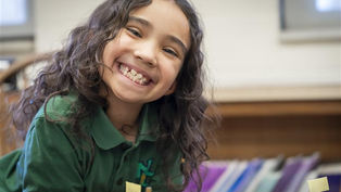 Smiling child with curly hair in a green shirt leans over a table with labeled mini flags. Classroom setting with colorful folders in background.
